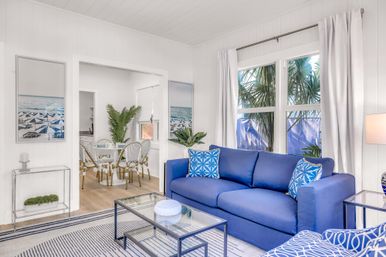 Bright coastal beach cottage living room with a bold blue sofa, patterned throw pillows, glass coffee table, dining nook, white walls, and palm trees visible through the window.