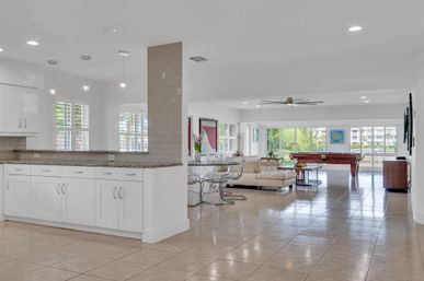 Bright open-concept coastal living area with white kitchen cabinets and granite counter, tiled floors, glass dining table, cream sectional, pool table, ceiling fan, and large windows framing palm trees and waterfront views.