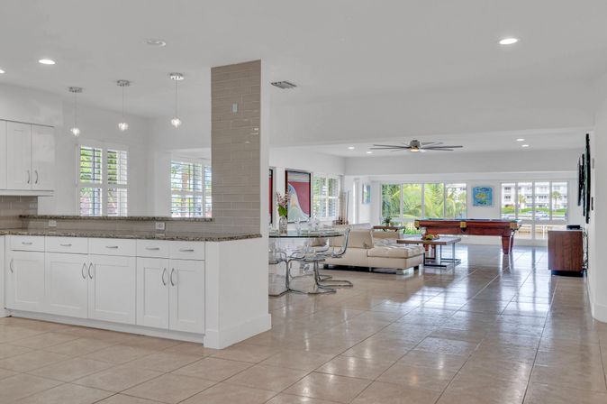Bright open-concept coastal living area with white kitchen cabinets and granite counter, tiled floors, glass dining table, cream sectional, pool table, ceiling fan, and large windows framing palm trees and waterfront views.