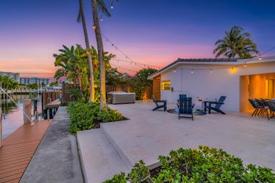 Waterfront sunset patio along a calm canal with palm trees and string lights — tiled outdoor lounge with Adirondack chairs around a fire pit, dining area, hot tub, and private dock.