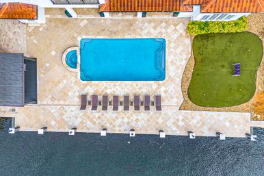 Aerial drone view of a waterfront patio: a rectangular blue pool with attached spa, row of lounge chairs on a tiled deck, adjacent putting green and dock.