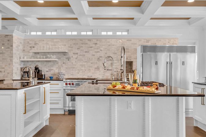 Bright modern luxury kitchen with white cabinetry and a dark-topped island, stainless steel refrigerator and range, light stone subway-tile backsplash, coffered ceiling, and a charcuterie board with champagne on the island.