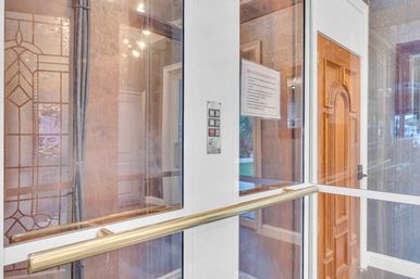 Glass-enclosed residential lobby vestibule with brass handrail, intercom panel, decorative stained-glass side window and carved wooden interior door with a posted notice.