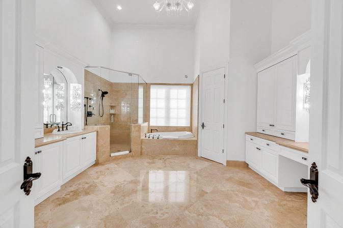 Spa-like luxury master bathroom with polished beige marble floors, glass walk-in shower, built-in soaking tub beneath a frosted window, white cabinetry with wood countertops, dual vanities and a crystal chandelier