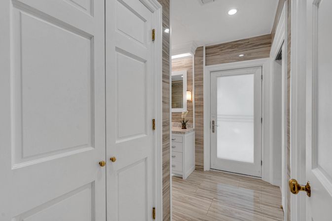 Bright modern residential bathroom entry with frosted glass door, white paneled closets and vanity, wood-look wall and floor tiles, recessed ceiling lights and brass hardware