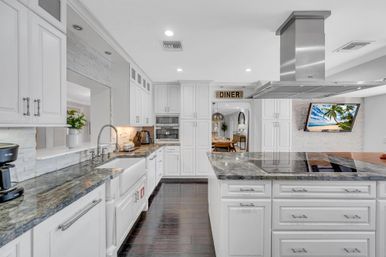 Bright open-concept white chef's kitchen with dark marble countertops and large island, stainless-steel range hood, farmhouse sink, dark hardwood floors, wall-mounted TV showing a beach scene and open view into a cozy dining area.
