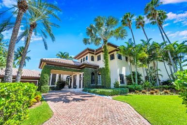 Mediterranean-style luxury villa with tile roof, ivy-covered entry, brick driveway and tall palm trees under a bright blue tropical sky