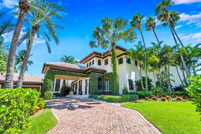 Mediterranean-style luxury villa with tile roof, ivy-covered entry, brick driveway and tall palm trees under a bright blue tropical sky