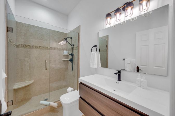 Contemporary home bathroom with frameless glass walk-in shower, beige tile and decorative border, matte-black shower fixtures, white countertop vanity with wood drawers, large mirror and industrial-style lights.