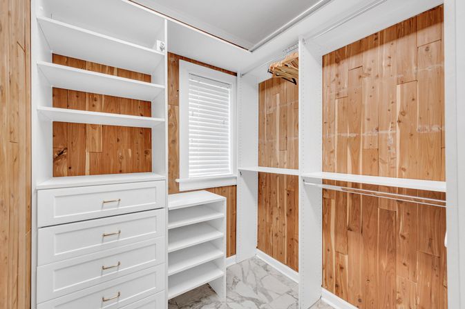 Bright walk-in closet with white built-in shelves and drawers, cedar wood-paneled walls, marble-look tile floor, small window with blinds, and hanging rods with wooden hangers.
