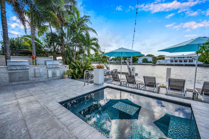 Waterfront modern pool and tiled patio with mosaic spa, built-in stainless outdoor kitchen, palm trees, blue umbrellas and lounge chairs along a calm canal.