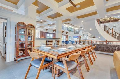 Bright open‑plan dining room with a long wooden table set for ten, mid‑century wooden chairs with navy cushions, coffered ceiling with gold insets, a modern linear pendant light, glass china cabinet, and a sweeping curved staircase
