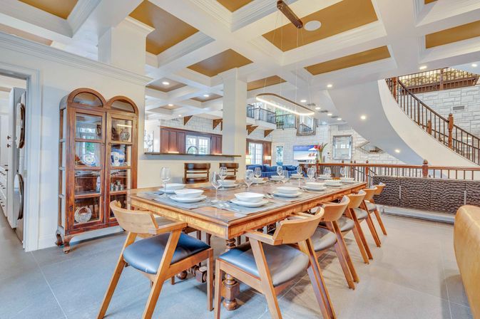 Bright open‑plan dining room with a long wooden table set for ten, mid‑century wooden chairs with navy cushions, coffered ceiling with gold insets, a modern linear pendant light, glass china cabinet, and a sweeping curved staircase