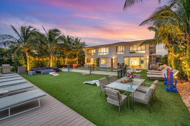 Tropical, resort-style luxury backyard at sunset — modern two-story home with lit palm trees, manicured lawn, outdoor dining table and lounge chairs, cornhole boards, sunken rectangular pool and string lights for al fresco entertaining.