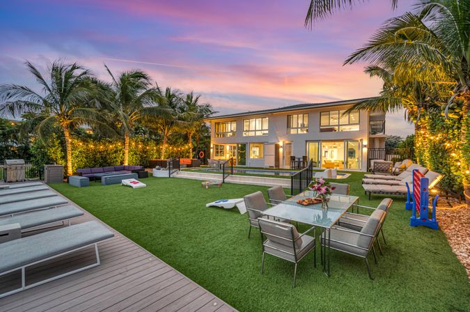 Tropical, resort-style luxury backyard at sunset — modern two-story home with lit palm trees, manicured lawn, outdoor dining table and lounge chairs, cornhole boards, sunken rectangular pool and string lights for al fresco entertaining.