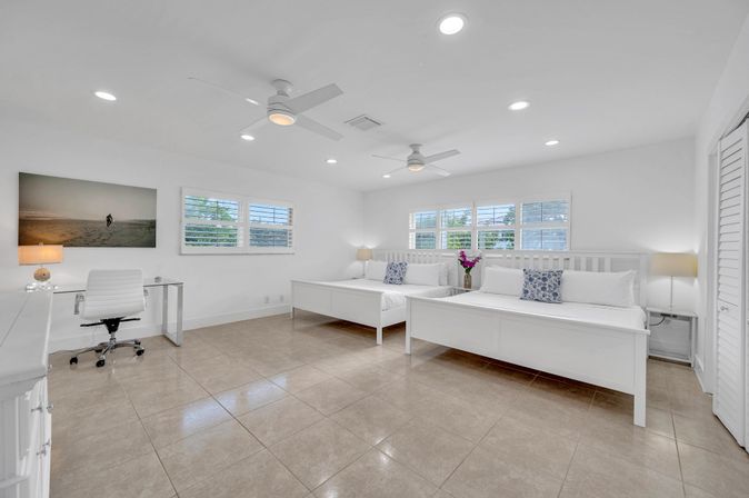 Airy, sunlit beach-style bedroom with two white platform beds, tile floor, ceiling fans, desk, bedside lamps and shuttered windows