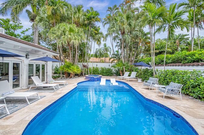 Tropical private backyard pool with bright blue water, raised circular spa, white lounge chairs and navy umbrellas on a stone patio, surrounded by tall palm trees and lush hedges.