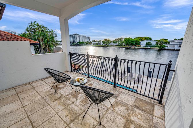 Waterfront balcony with tiled floor, two black woven chairs and small table with coffee and breakfast, wrought-iron railing overlooking a calm canal lined with palm trees and low-rise waterfront buildings under a blue sky.