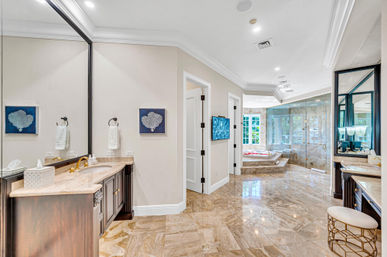 Spacious spa-like master bathroom with polished marble floors, dual dark-wood vanities with gold faucets, oversized mirrors, frameless glass shower, and sunken jetted tub by bay windows