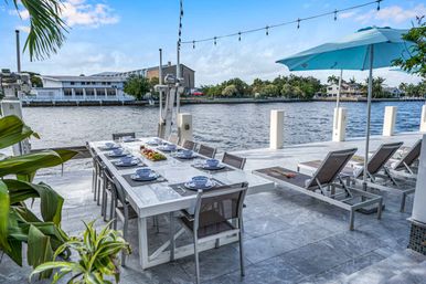 Inviting canal-side patio with a marble dining table set for eight, blue place settings, tropical plants, string lights and lounge chairs under a turquoise umbrella