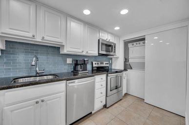 Bright modern kitchen with white cabinets, blue subway tile backsplash, dark granite countertops, stainless steel sink, dishwasher, range and microwave, plus a stacked washer-dryer in a closet.