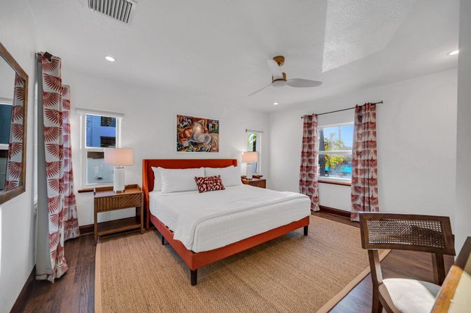 Bright coastal resort bedroom with king bed and coral headboard, crisp white linens, patterned tropical curtains, wooden nightstands and ceiling fan, with palm-tree and pool view through windows.