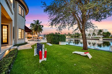 Sunset-lit tropical waterfront backyard with palm trees, manicured lawn, oversized Connect Four and cornhole games, private dock and canal beside a luxury home.