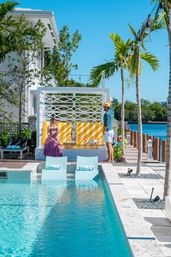 Sunlit tropical waterfront pool scene with turquoise in-pool loungers, a yellow-striped cabana, palm trees and string lights; two people chatting by a dock overlooking a calm blue bay at a coastal resort.