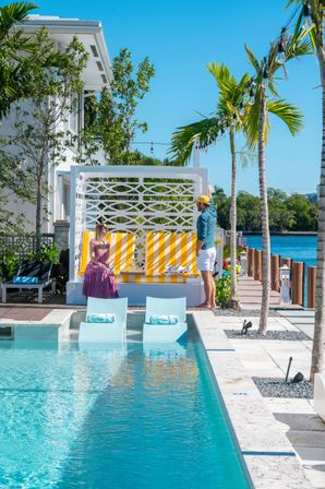 Sunlit tropical waterfront pool scene with turquoise in-pool loungers, a yellow-striped cabana, palm trees and string lights; two people chatting by a dock overlooking a calm blue bay at a coastal resort.