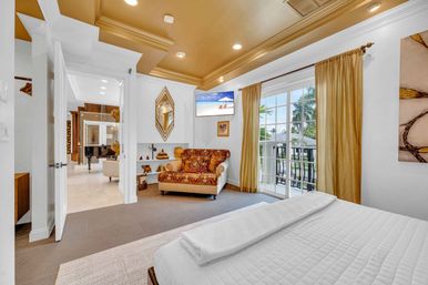 Sunlit tropical master bedroom with white quilted bed, patterned loveseat, gold tray ceiling and curtains, glass doors opening to a balcony with palm trees and water view.