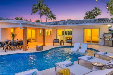 Inviting tropical backyard at sunset with a modern single-story home, illuminated patio, in-ground swimming pool with lounge chairs and in-pool loungers, outdoor dining area, palm trees and a gas grill.