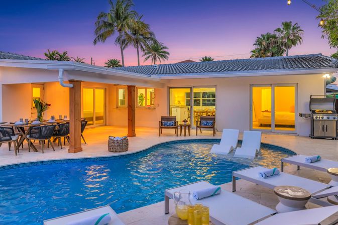 Inviting tropical backyard at sunset with a modern single-story home, illuminated patio, in-ground swimming pool with lounge chairs and in-pool loungers, outdoor dining area, palm trees and a gas grill.