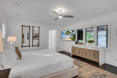 Sunlit coastal-style master bedroom with white bedding on a low platform bed, ceiling fan, wall-mounted TV showing a tropical beach, hardwood floors, built-in wood-and-white dresser and wide windows with blinds