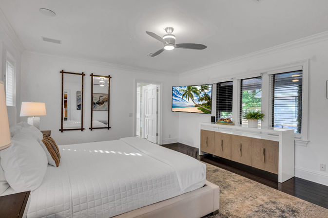 Sunlit coastal-style master bedroom with white bedding on a low platform bed, ceiling fan, wall-mounted TV showing a tropical beach, hardwood floors, built-in wood-and-white dresser and wide windows with blinds