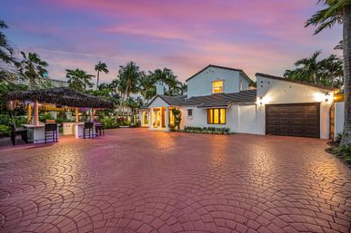 Mediterranean-style white stucco home with tiled roof, wide patterned red driveway, tiki thatched outdoor bar, swaying palm trees and a pink sunset sky.