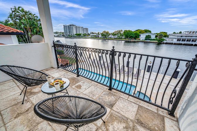 Waterfront balcony with two black woven lounge chairs and a small glass table holding coffee and fruit, ornate iron railing overlooking a pool and calm river with low-rise waterfront buildings and palm trees under a blue sky.