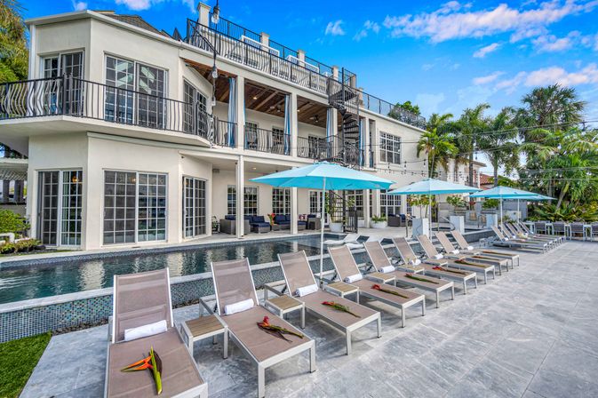 Sun-soaked tropical pool deck at a luxury waterfront villa with rows of loungers and turquoise umbrellas beside a narrow lap pool, palm trees and balcony terraces