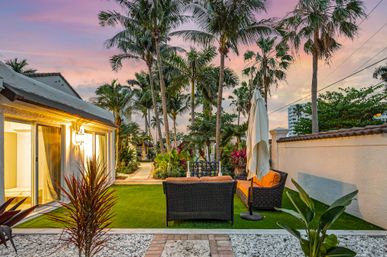 Cozy tropical backyard patio at sunset — wicker sofa with orange cushions, closed umbrella, manicured lawn, palm trees and a stucco home with sliding glass door.
