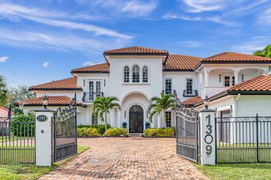 Gated Mediterranean-style two-story villa with white stucco, terracotta roof, arched double-door entry, palm trees and a brick driveway under a bright blue sky.