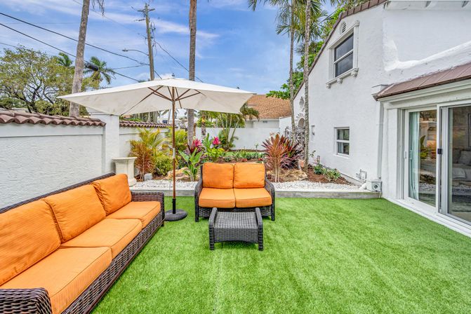 Sunny tropical backyard patio with white stucco house, green artificial turf, wicker sofas with bright orange cushions, large white umbrella, lush palms and sliding glass doors.