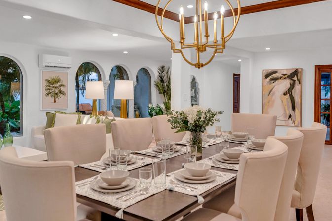 Sunlit coastal villa dining room with a long wooden table set for eight, beige upholstered chairs, floral centerpiece, gold chandelier, and arched windows framing palm trees.