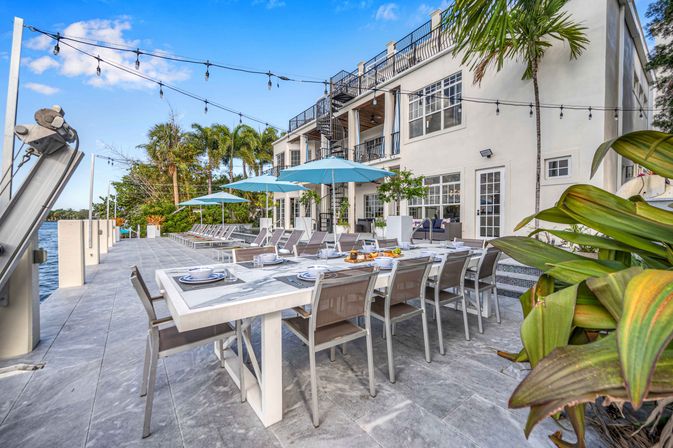 Sunny waterfront patio with a long outdoor dining table set with plates and snacks, blue umbrellas and lounge chairs along a dock, string lights and palm trees beside a white coastal villa
