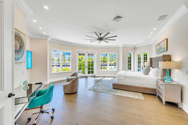 Bright, spacious waterfront master bedroom with king bed and tufted headboard, turquoise accents, glass desk, cozy seating, hardwood floors, ceiling fan and French doors opening to a sunny canal view.