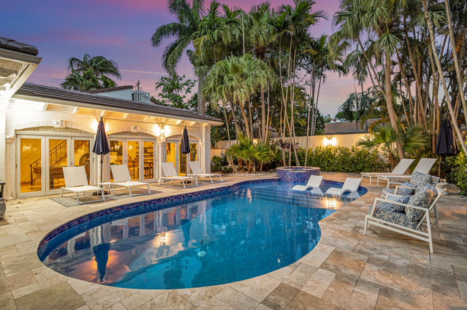 Tropical backyard swimming pool at dusk with curved blue-tiled pool and raised spa, stone tile patio with white lounge chairs, umbrellas and patterned cushions, surrounded by tall palm trees and warm outdoor lighting.