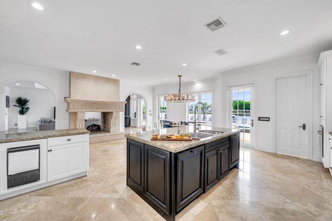 Sunlit open-concept luxury kitchen with a large dark island and granite countertops, crystal chandelier, ornate stone fireplace, polished marble floors, and glass doors opening to a waterfront patio.