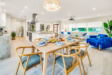 Open-concept modern kitchen, dining and living area with a wooden dining table set for six, marble-topped island and stainless steel range hood, blue velvet accent chairs, light wood floors and large windows with garden views.