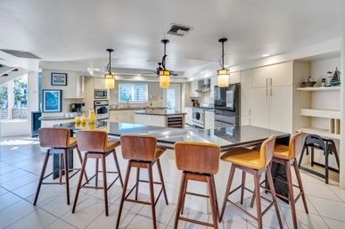 Bright open-concept kitchen featuring a large granite island with five mid-century wooden bar stools, pendant lights, stainless steel appliances, white cabinetry, tiled floor, and sunlit windows.