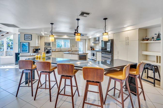 Bright open-concept kitchen featuring a large granite island with five mid-century wooden bar stools, pendant lights, stainless steel appliances, white cabinetry, tiled floor, and sunlit windows.