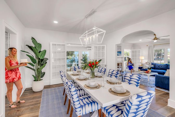 Bright coastal-style open-plan dining and living area with white rectangular table set for eight, blue-and-white patterned chairs, geometric pendant light and floral centerpiece; hardwood floors, a woman in a red dress carrying a cake, and another person relaxing on a blue sofa in the adjacent living room.