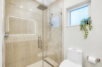 Bright modern bathroom with glass walk-in shower, wood-effect wall paneling and hexagon tile, rainfall showerhead, white toilet and small window with trailing plant